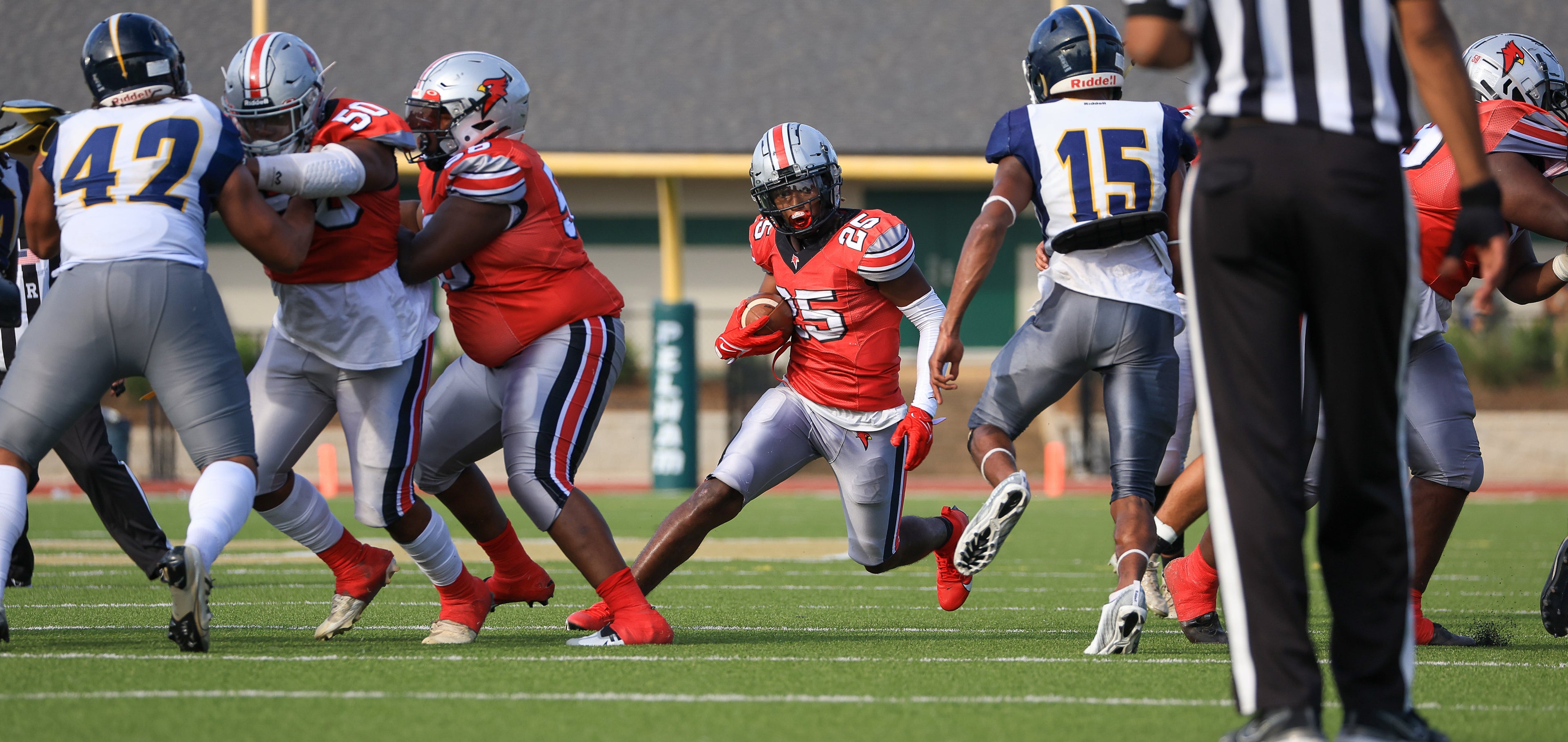 American football player in red jersey running with ball, surrounded by opponents and referee on field